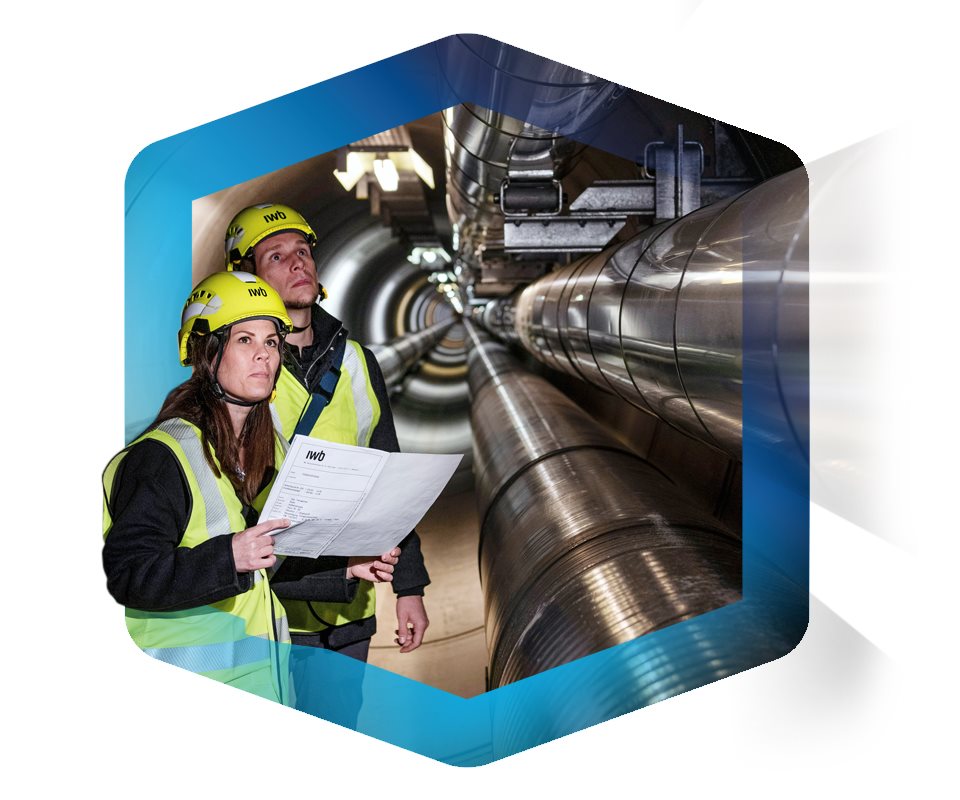 Zwei IWB-Mitarbeitende in Schutzkleidung und Helmen inspizieren ein grosses unterirdisches Leitungssystem. Sie halten Pläne in der Hand und stehen in einem beleuchteten Tunnel mit grossen Rohren – Symbolbild für Versorgungssicherheit und Infrastruktur.
